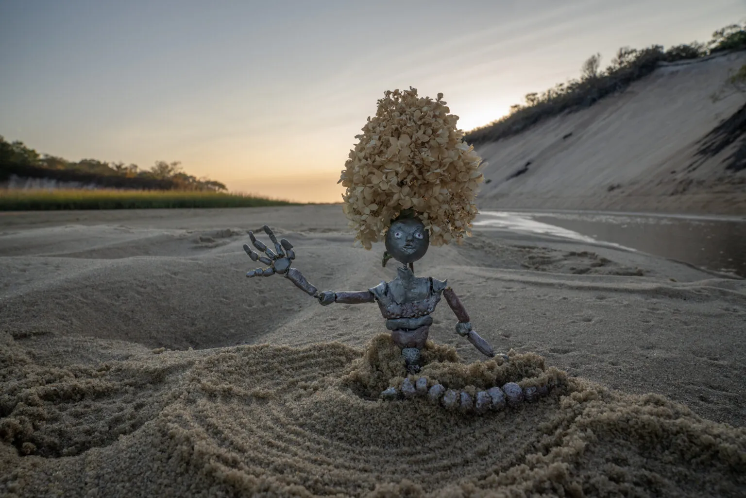 Metallic humanoid half-buried in sand with yellow leafy hair on top of its head