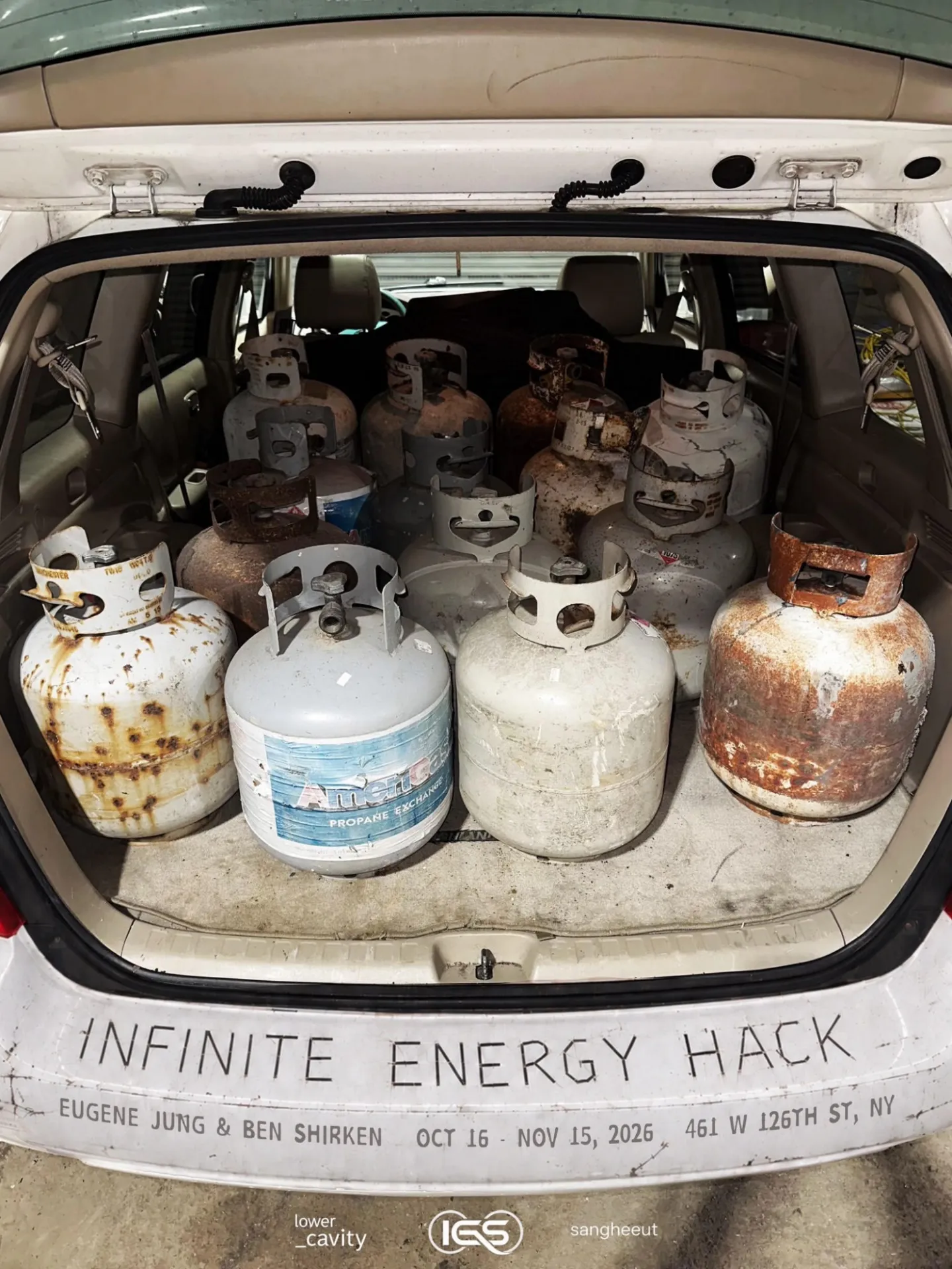 Dilapidated propane canisters in what appears to be the trunk of a mini van. On the bumper, "INFINITE ENERGY HACK" is written in all caps and, in smaller type, the event details.
